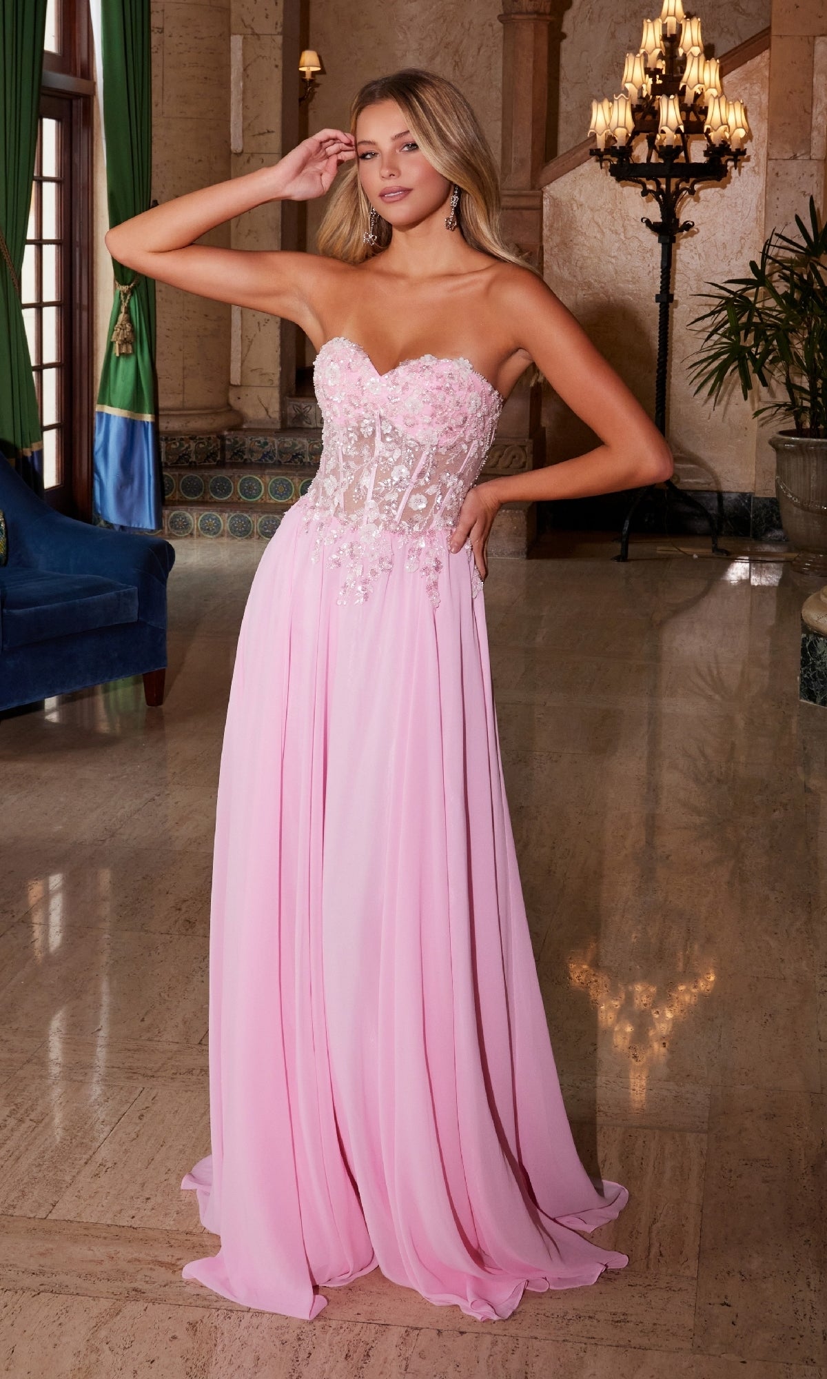 A woman stands indoors wearing the Nox Anabel C1617, a strapless sweetheart prom dress in pink, featuring a sheer embellished bodice and a flowing floor-length skirt.
